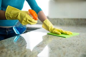 Parent cleaning a kitchen table with a spray bottle as young children and a dog play nearby in a sunlit room.