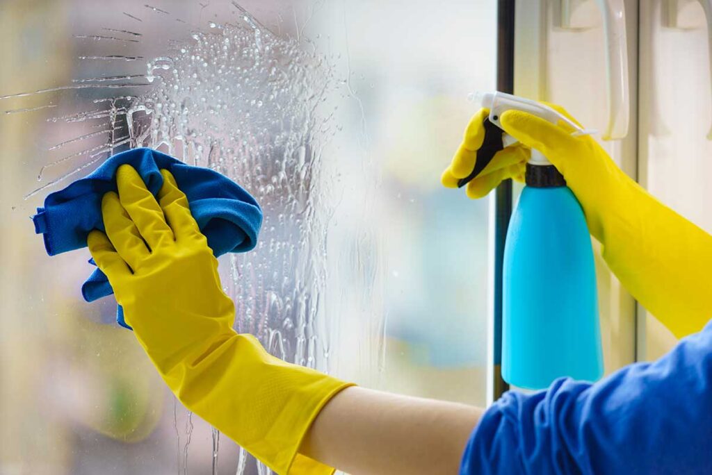 A person in casual clothing sprays a countertop with a clear bottle labeled "eco-friendly cleaner" next to a potted plant.